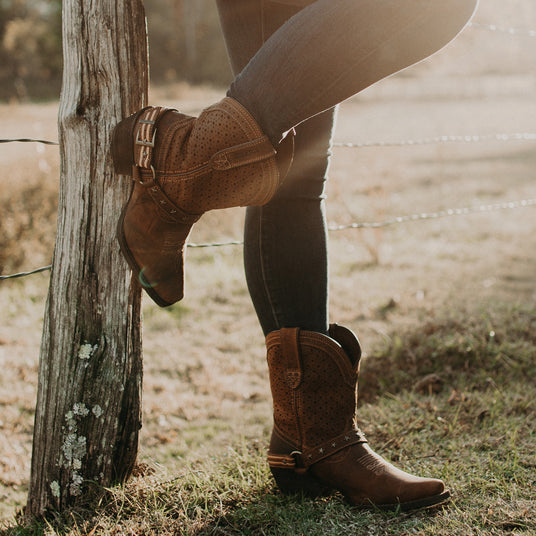 Close up image of a woman wearing a pair of Durango Crush boots, resting one foot vertically on a wooden post